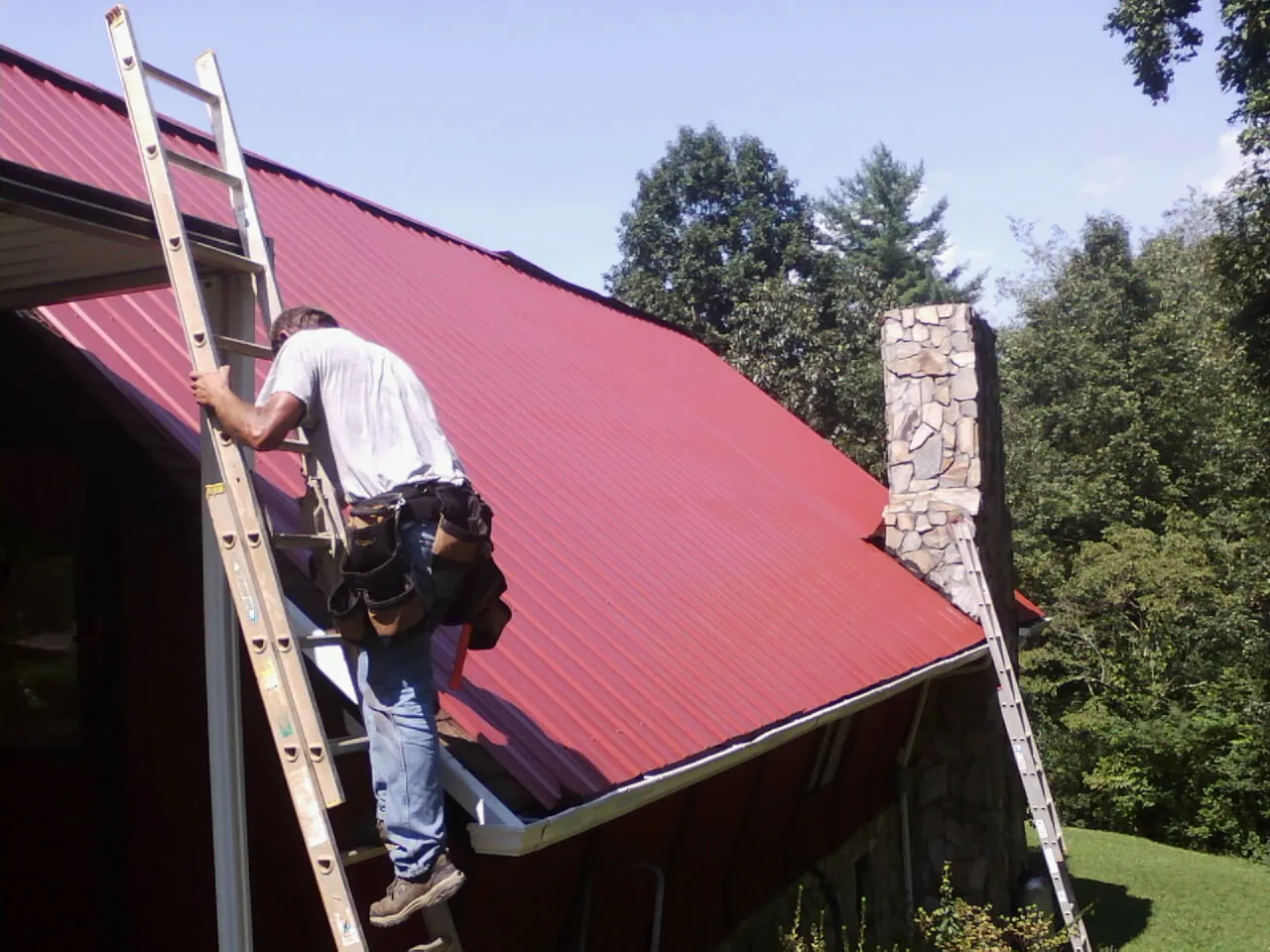 Roofer on a ladder beside a red metal roof and stone chimney — Soffit & Fascia Repair by Roofing Brothers in Valley Center